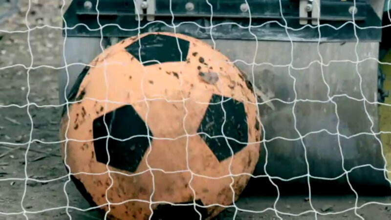 An oversized, salmon-coloured football being pushed by a steamroller into a goal. An oversized, salmon-coloured football being pushed by a steamroller into a goal.
