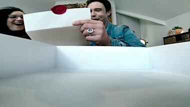 A shot from inside a shallow white cardboard box. Over the box are Ed Gamble and Rose Matafeo reading a task. Rose appears to be laughing.
