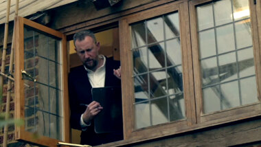 Alex Horne leaning slightly out an upstairs window. He is looking down towards the ground and holding a sealed task.