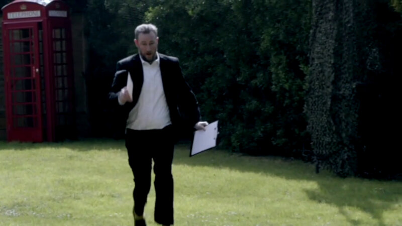 Alex Horne running from a red phone box towards the camera across the Taskmaster house lawn. He is holding a sealed task in one hand, and a clipboard in the other. Alex Horne running from a red phone box towards the camera across the Taskmaster house lawn. He is holding a sealed task in one hand, and a clipboard in the other.