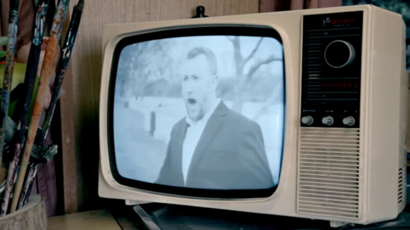 A close-up of a tan plastic table top CRT television. Showing on the TV is a picture Alex Horne with his mouth open as if speaking or singing. The picture is in black and white. A close-up of a tan plastic table top CRT television. Showing on the TV is a picture Alex Horne with his mouth open as if speaking or singing. The picture is in black and white.