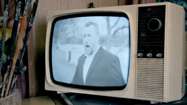 A close-up of a tan plastic table top CRT television. Showing on the TV is a picture Alex Horne with his mouth open as if speaking or singing. The picture is in black and white.