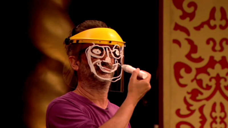 David Baddiel on the Taskmaster studio stage. He is wearing a yellow visor with a clear plastic shield covering his face. He is drawing a face over his face using a white paint pen. David Baddiel on the Taskmaster studio stage. He is wearing a yellow visor with a clear plastic shield covering his face. He is drawing a face over his face using a white paint pen.