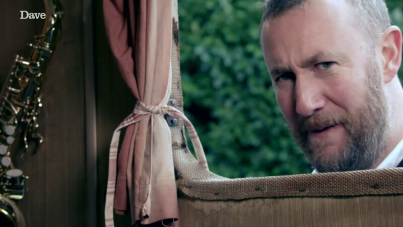 Alex Horne looking at the camera through the caravan window. He is looking perplexed. A portion of the pink curtains can be seen as well as part of a saxophone. Alex Horne looking at the camera through the caravan window. He is looking perplexed. A portion of the pink curtains can be seen as well as part of a saxophone.