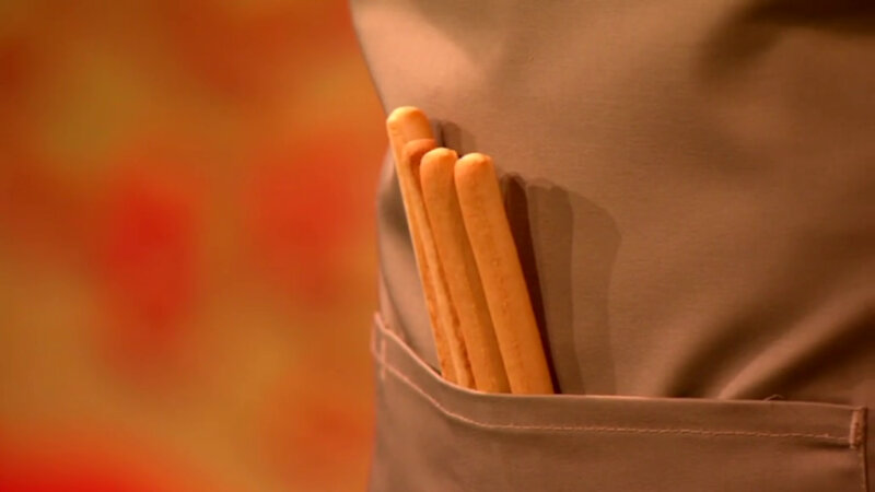 A close-up of four skinny breadsticks sticking out of a tan apron pocket. A close-up of four skinny breadsticks sticking out of a tan apron pocket.