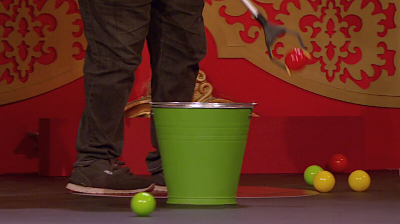 A close-up of Johnny Vegas' legs on the Taskmaster stage. He is picking up a small red ball with a grabbing tool. In front of him is a green bucket as well as two green, two yellow, and another red ball. A close-up of Johnny Vegas' legs on the Taskmaster stage. He is picking up a small red ball with a grabbing tool. In front of him is a green bucket as well as two green, two yellow, and another red ball.