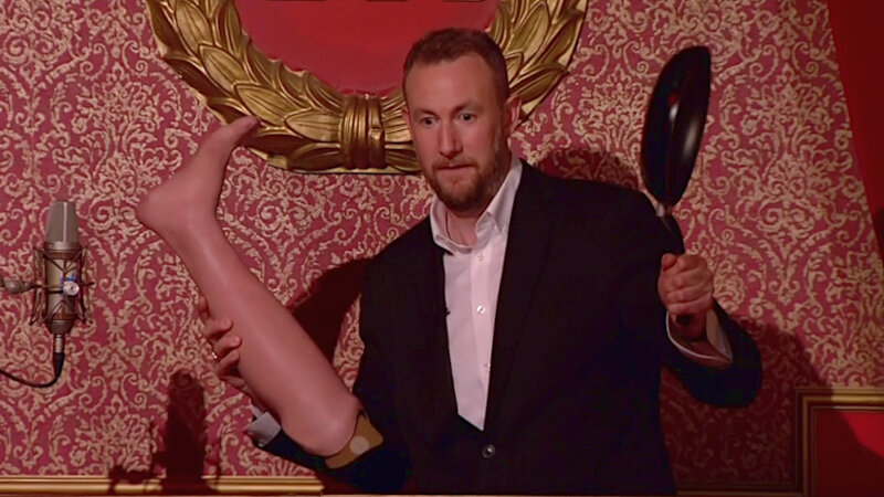 Alex Horne on the Taskmaster studio stage. He is next to a microphone and is holding a prosthetic leg and a frying pan. Alex Horne on the Taskmaster studio stage. He is next to a microphone and is holding a prosthetic leg and a frying pan.