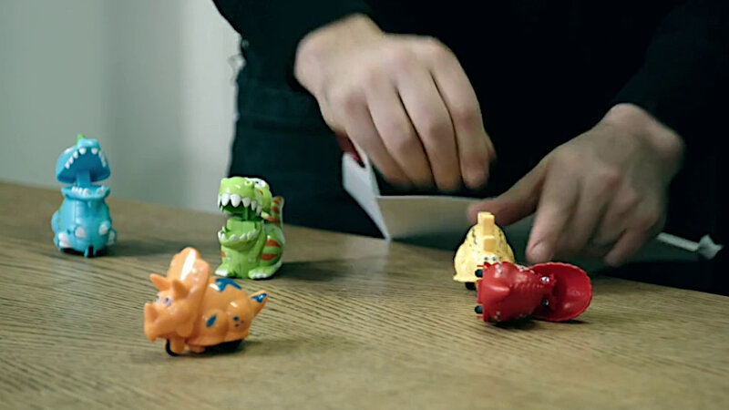 A close-up of Richard Herring's hands opening a task. In front of him on a wooden table are five wind-up toy dinosaurs in blue, green, orange, yellow, and red. A close-up of Richard Herring's hands opening a task. In front of him on a wooden table are five wind-up toy dinosaurs in blue, green, orange, yellow, and red.