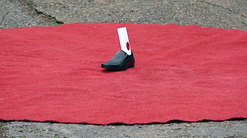 A round red rug on concrete outside the Taskmaster house. In the centre of the rug is a black men's shoe with a sealed task inside. A round red rug on concrete outside the Taskmaster house. In the centre of the rug is a black men's shoe with a sealed task inside.