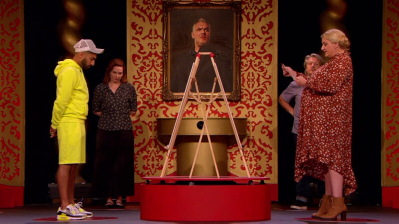 Daisy May Cooper, Katherine Parkinson, Mawaan Rizwan and Richard Herring standing on the Taskmaster studio stage. They are surrounding an artist's easel on a red rotating platform. Daisy May is reading a task. Daisy May Cooper, Katherine Parkinson, Mawaan Rizwan and Richard Herring standing on the Taskmaster studio stage. They are surrounding an artist's easel on a red rotating platform. Daisy May is reading a task.