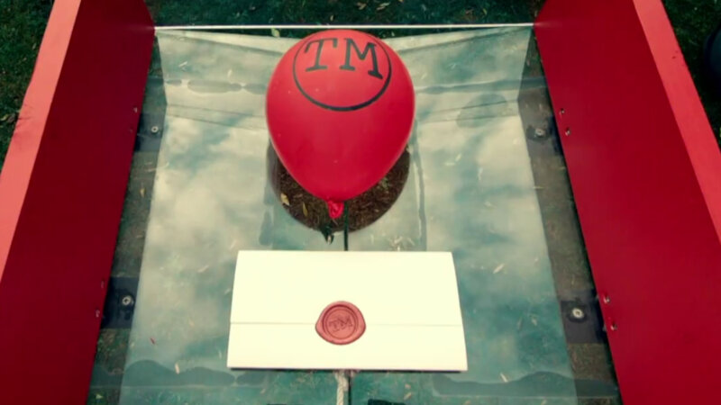 A close-up of a red balloon with the Taskmaster logo. It is resting inside a red wooden frame with a clear plexiglass bottom. Beside the balloon is a sealed task. A close-up of a red balloon with the Taskmaster logo. It is resting inside a red wooden frame with a clear plexiglass bottom. Beside the balloon is a sealed task.