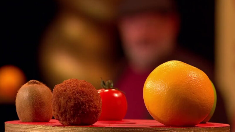 A close-up of a number of round edible food items on top of a wood stand. They are a scotch egg, a kiwi fruit, a tomato, a green apple and an orange. Johnny Vegas can be seen out of focus in the background. A close-up of a number of round edible food items on top of a wood stand. They are a scotch egg, a kiwi fruit, a tomato, a green apple and an orange. Johnny Vegas can be seen out of focus in the background.