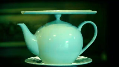 A sealed task resting on a teapot resting on a small plate. All the items are white but appear green due to the lighting.
