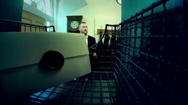 A shot from inside a black wire basket in the Taskmaster house foyer. Inside the basket is a sealed task. Alex Horne can be seen in the background.