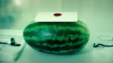 A close-up of a watermelon resting on a white table in the Taskmaster house lab. On top of the watermelon is a sealed task. The watermelon is in between two pairs of disposable white plastic coveralls and two pairs of safety goggles.