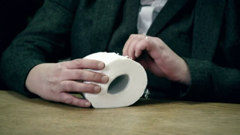 A close-up of Johnny Vegas' hand peeling apart a toilet roll.
