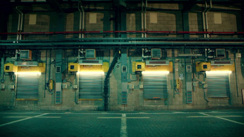 A large room with a brick floor and a concrete wall. Along the wall are garage style lifting doors in corrugated metal. Around the doors are many electrical panels, pipes, wired conduits and other mechanical flotsam. A large room with a brick floor and a concrete wall. Along the wall are garage style lifting doors in corrugated metal. Around the doors are many electrical panels, pipes, wired conduits and other mechanical flotsam.