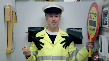 A middle-aged white man with salt and pepper hair and beard standing in a white cubicle. He is wearing a yellow jacket with silver reflector tape. A white captain's style hat with a black bill. In one hand he is holding a small yellow rubber duck and in the other a round yellow and red sign that reads 'stop' and 'children' in black script. On one side of the cubicle hang a number of framed pieces of art, and a copper desert mould. On the other side is a yellow capital letter 'T'. behind him there are two holes in the wall of cubicle. Black gloved hands are reaching through and resting on the man's chest.
