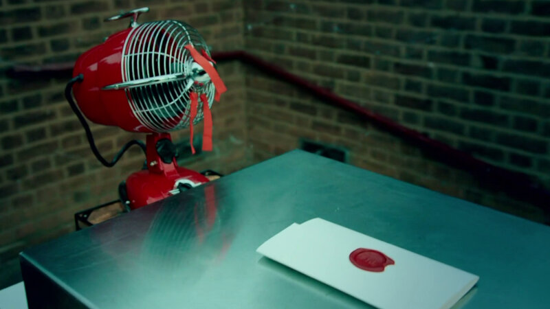 A metal box resting on a table next to the Taskmaster house. Next to the box is a red electric fan with orange ribbons tied to the mesh blade cover. On top of the box is a sealed task. A metal box resting on a table next to the Taskmaster house. Next to the box is a red electric fan with orange ribbons tied to the mesh blade cover. On top of the box is a sealed task.