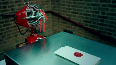 A metal box resting on a table next to the Taskmaster house. Next to the box is a red electric fan with orange ribbons tied to the mesh blade cover. On top of the box is a sealed task.