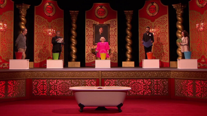 A long shot of the Taskmaster studio stage. In front of the stage is a claw foot bathtub. On the stage are the five contestants standing in a semi-circle in the same order they sat on their stage chairs. Next to each contestant is a white wooden box and a coloured tin bucket. From left they are red, green, yellow, orange, and blue. A long shot of the Taskmaster studio stage. In front of the stage is a claw foot bathtub. On the stage are the five contestants standing in a semi-circle in the same order they sat on their stage chairs. Next to each contestant is a white wooden box and a coloured tin bucket. From left they are red, green, yellow, orange, and blue.