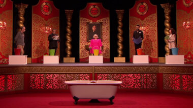 A long shot of the Taskmaster studio stage. In front of the stage is a claw foot bathtub. On the stage are the five contestants standing in a semi-circle in the same order they sat on their stage chairs. Next to each contestant is a white wooden box and a coloured tin bucket. From left they are red, green, yellow, orange, and blue.
