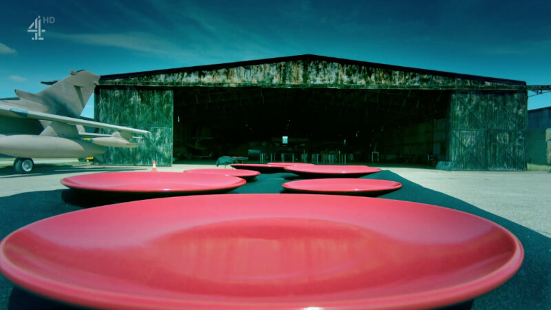 A low shot of a table with a black tablecloth covered in red plates. The table is in front of a rusty and dirty aeroplane hanger. Part of a tan aeroplane can be seen. A low shot of a table with a black tablecloth covered in red plates. The table is in front of a rusty and dirty aeroplane hanger. Part of a tan aeroplane can be seen.