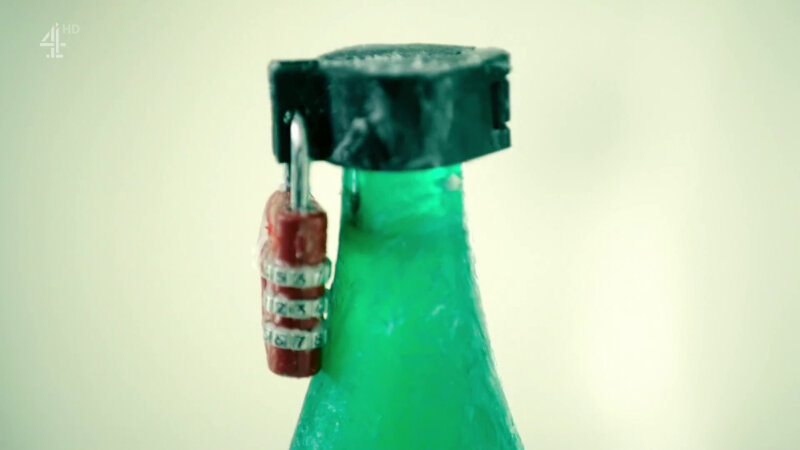 A close-up of a green glass bottle coated in Vaseline. It is sealed with a metal clamp held together with a red padlock. A close-up of a green glass bottle coated in Vaseline. It is sealed with a metal clamp held together with a red padlock.