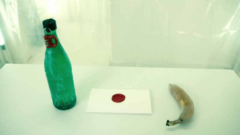 The table in the Taskmaster house lab. On it is a green bottle coated in Vaseline. It is sealed with a metal clamp held together with a red padlock. Next to the bottle is a sealed task and next to that a frozen banana. The table in the Taskmaster house lab. On it is a green bottle coated in Vaseline. It is sealed with a metal clamp held together with a red padlock. Next to the bottle is a sealed task and next to that a frozen banana.
