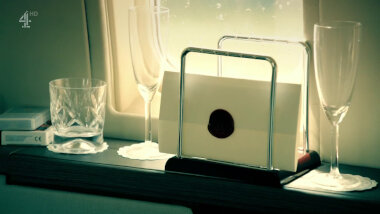 A sealed task brief on a shelf by an aeroplane's window. Two champagne flutes and a whiskey glass sit empty on the ledge near it as well as some playing cards.