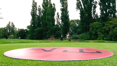 A red circle with the letters TM lies flat on manicured grass. In the distance, Sarah Kendall sits on a lifeguard's chair. Alex Horne stands near her. Charlotte Ritchie and Jamali Maddox are visible in the distance.