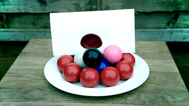A sealed task brief is set up on a plate full of snooker balls on a wooden table.
