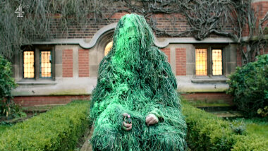 A person in a ghillie suit sits in verdant courtyard.