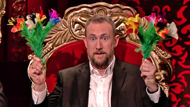 Alex Horne in his throne holding two magician's bouquets.