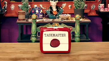 A plastic lunch box on the living room table. Where you'd normally see a character or design on a lunchbox, the design is of a sealed task brief and the word 'Taskmaster'.
