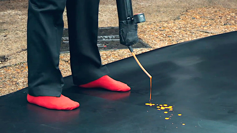 Alex Horne stands on a black mat in the driveway. He's wearing red socks. A tube hanging down from him is slowly dripping yellow paint on to the black mat. Alex Horne stands on a black mat in the driveway. He's wearing red socks. A tube hanging down from him is slowly dripping yellow paint on to the black mat.