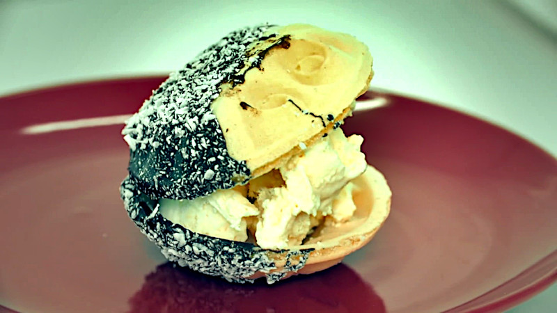 A dessert on a red plate. Off-white ice cream sandwiched between two bowl shaped wafers half dipped in what looks like chocolate and coconut flakes. A dessert on a red plate. Off-white ice cream sandwiched between two bowl shaped wafers half dipped in what looks like chocolate and coconut flakes.
