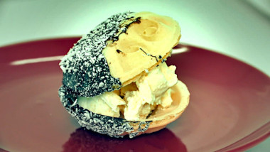 A dessert on a red plate. Off-white ice cream sandwiched between two bowl shaped wafers half dipped in what looks like chocolate and coconut flakes.