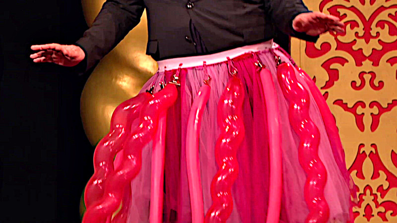 A contestant on the studio stage is wearing a pink tutu with several inflated tube-shaped balloons attached to the waist of the tutu. A contestant on the studio stage is wearing a pink tutu with several inflated tube-shaped balloons attached to the waist of the tutu.