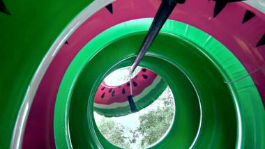 View from the centre of a stack of inflated inner tubes. They're coloured pink and green like watermelon. A long stick also goes through the centre of the stack and is piercing the centre of one more innertube hanging in the air.