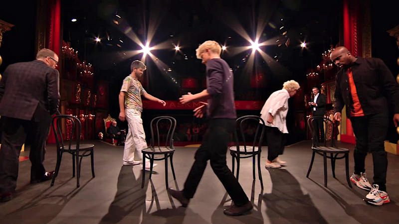 The cast of UK series 16 on the studio stage walk in a circle around four black chairs. The cast of UK series 16 on the studio stage walk in a circle around four black chairs.