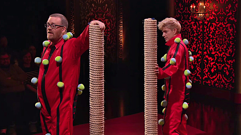 Mae Martin and Frankie Boyle on the studio stage. They're each wearing a red boiler suit with Velcro stripes and cloth balls stuck to the stripes. They're each holding onto a rope-covered scratching post roughly their own height. Mae Martin and Frankie Boyle on the studio stage. They're each wearing a red boiler suit with Velcro stripes and cloth balls stuck to the stripes. They're each holding onto a rope-covered scratching post roughly their own height.