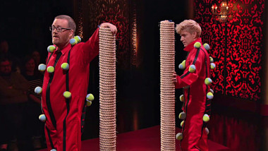 Mae Martin and Frankie Boyle on the studio stage. They're each wearing a red boiler suit with Velcro stripes and cloth balls stuck to the stripes. They're each holding onto a rope-covered scratching post roughly their own height.