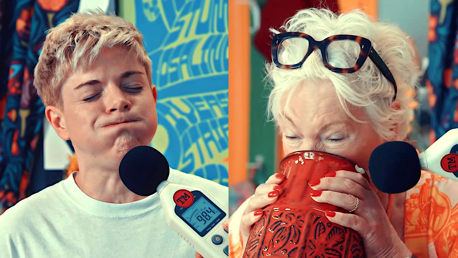 Mae Martin and Jenny Eclair in split-screen. Mae's lips are closed and cheeks are puffed out as Alex holds a decibel meter to their chin. Jenny's mouth and nose are in a red vase she's holding up to her face. A decibel meter is being held near the lip of the vase. Mae Martin and Jenny Eclair in split-screen. Mae's lips are closed and cheeks are puffed out as Alex holds a decibel meter to their chin. Jenny's mouth and nose are in a red vase she's holding up to her face. A decibel meter is being held near the lip of the vase.