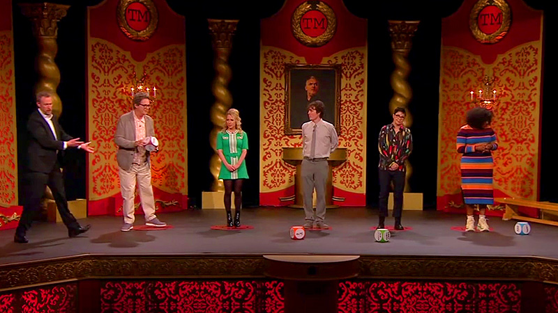 The cast of Taskmaster UK series 16 on the studio stage. There are three large novelty dice on the floor. Alex Horne gestures toward Julian Clary who is about to throw the die he is holding.
