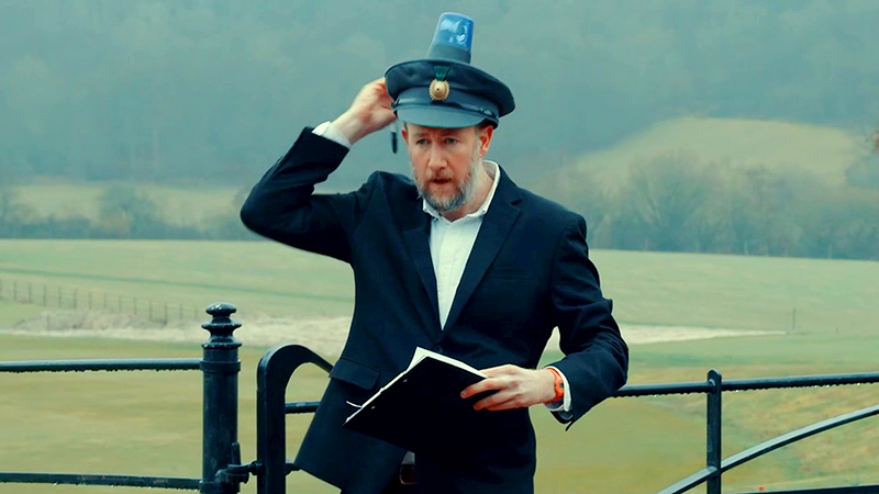 Alex Horne standing in front of a fence. He's wearing a policeman's hat with a siren on top of it, holding a clipboard and pen. Alex Horne standing in front of a fence. He's wearing a policeman's hat with a siren on top of it, holding a clipboard and pen.