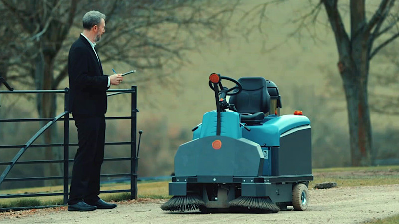Alex Horne standing outdoors looking at a small street sweeper. Alex Horne standing outdoors looking at a small street sweeper.