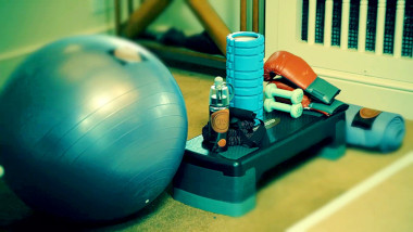 A pile of exercise equipment on carpet: an exercise ball, a step, weights, a water bottle, a roller, boxing gloves and a yoga mat.