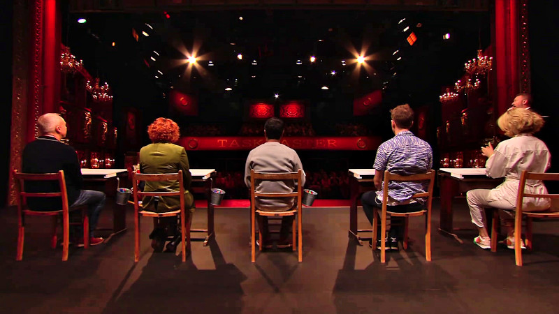 The cast of Taskmaster UK series 17 on the studio stage viewed from behind seated at desks. There's a pail of coloured markers attached to the side of each desk. The cast of Taskmaster UK series 17 on the studio stage viewed from behind seated at desks. There's a pail of coloured markers attached to the side of each desk.