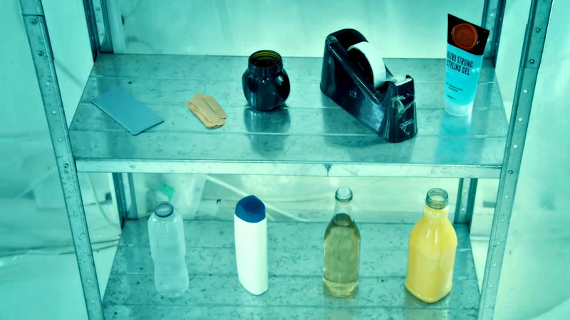 Metal shelves lined with items: blu tack, plasters, Marmite, tape, styling gel, and four plastic bottles with various coloured liquids inside. Metal shelves lined with items: blu tack, plasters, Marmite, tape, styling gel, and four plastic bottles with various coloured liquids inside.